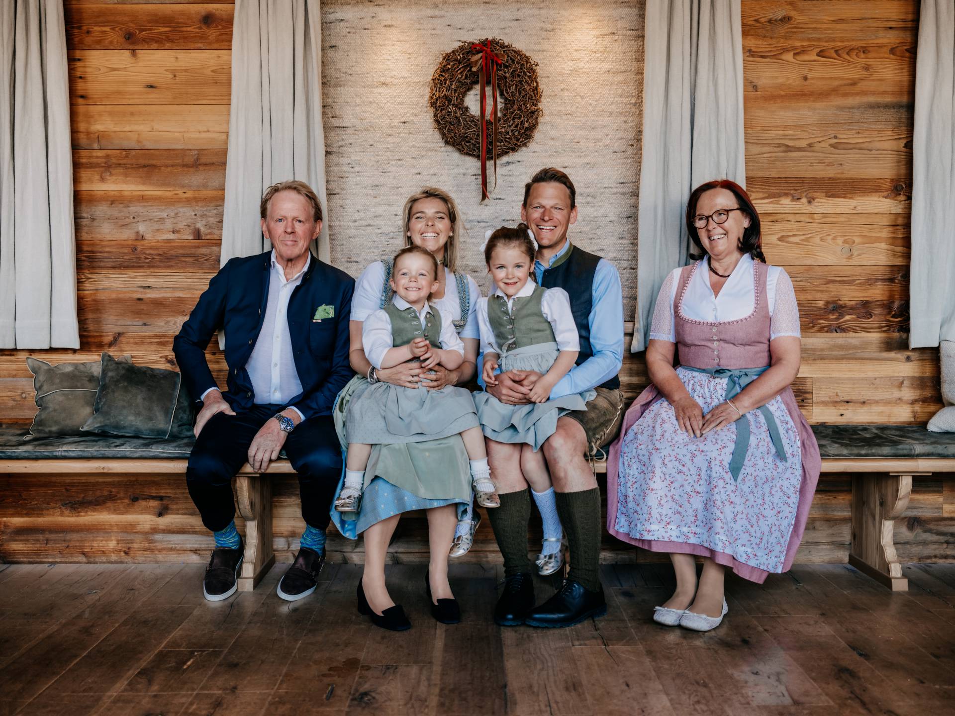 Three generations of a family dressed in traditional Alpine attire sit smiling on a wooden bench in a cozy, wood-paneled room. A decorative wreath hangs on the wall in the background. The family atmosphere and alpine decor convey warmth, hospitality, and a strong connection to the region.